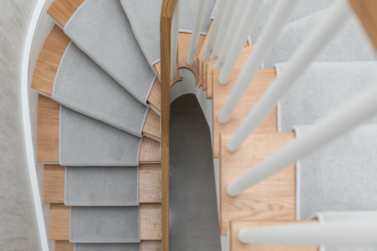 Overhead view down a curving u shaped wooden staircase with grey carpet runners, white balusters and a curved handrail